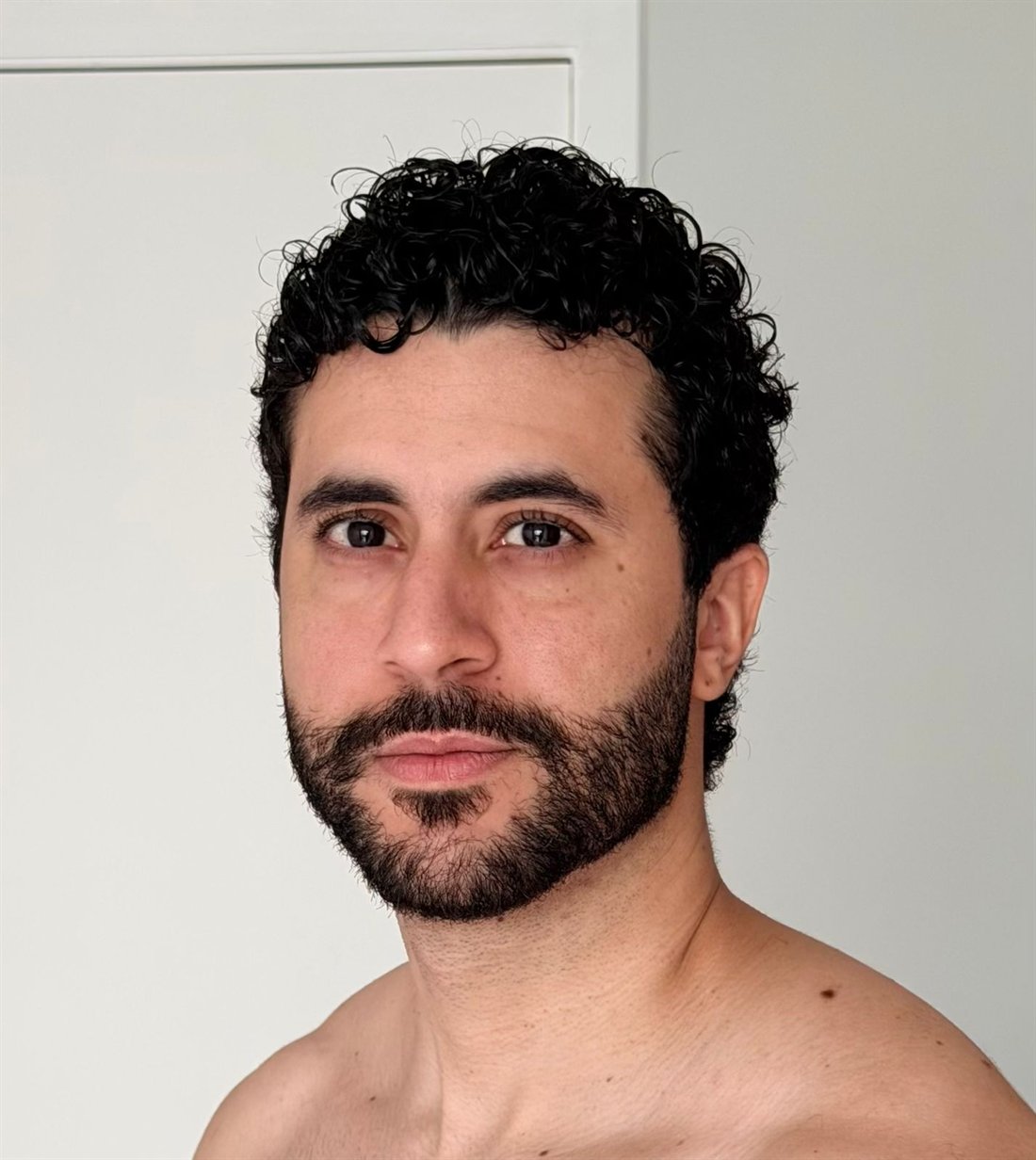Foto de celular, sem tratamento, de um homem de barba e cabelo cacheado contra parede branca